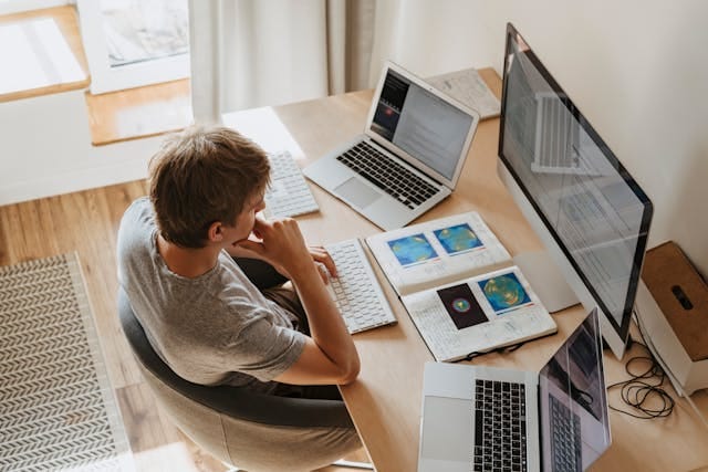 An adult self-learner studying at a desk with multiple screens and printed reference materials